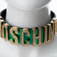 A Moschino letter necklace displayed on a white mannequin bust. The necklace features gold-tone metal letters with rhinestone accents, set against a vibrant green background with a black inner lining.