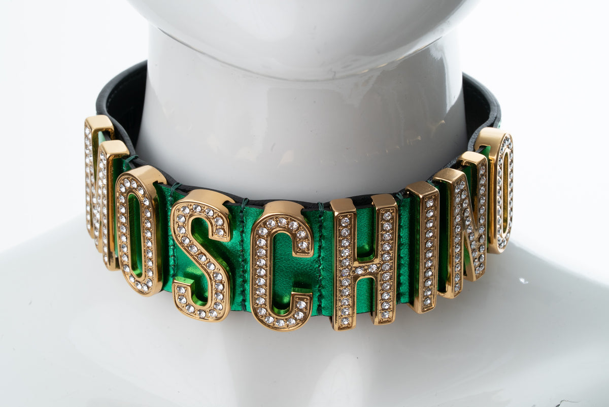 A Moschino letter necklace displayed on a white mannequin bust. The necklace features gold-tone metal letters with rhinestone accents, set against a vibrant green background with a black inner lining.